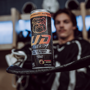 A can of UD Energy performance drink in Strawberry Lemonade flavor is in sharp focus, resting on a hockey stick's blade. In the blurred background, a hockey player in a locker room setting appears relaxed. The can's design, featuring a prominent bulldog logo and the slogan "Fuel Your Passion, Feed Your Beast" and "No Preservatives," targets athletes who seek a healthy, energizing boost with 249mg of caffeine and only 45 calories.