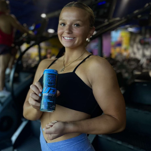A smiling woman in workout attire holding a can of UD Energy Blue Raspberry flavor in a gym setting. The can&